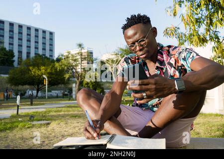 success is in the organization, young man of african ethnicity with glasses and short hair, is sitting outdoors using the phone and making notes in hi Stock Photo