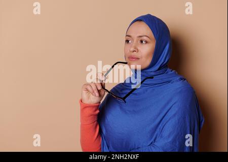 Confident Arab Muslim woman in blue hijab, holding eyeglasses and looking away thoughtfully on isolated beige background Stock Photo