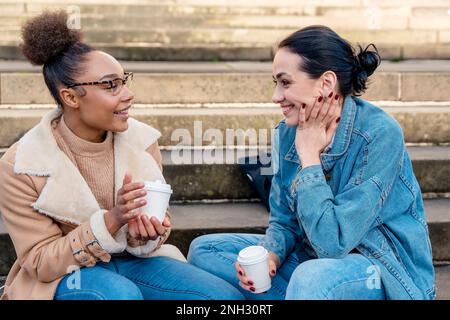 two student women in denim jackets are talking to each other, on the phone, laughing, and drinking coffee on stairs in the city.  social media divides Stock Photo