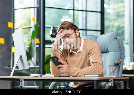 A young male programmer, freelancer, businessman, office worker sits at the table and looks at the phone. Worriedly holding his head, received bad news, message, chats. Stock Photo