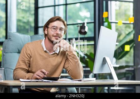 Portrait of a young man in glasses working at a computer in a modern office. He looks at the camera, smiles. Stock Photo
