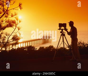 Silhouette of a man photographer, using a 4x5 camera on a tripod taking a picture of the sunset out over the ocean Stock Photo