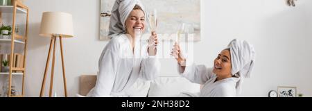joyful multiethnic women in white bathrobes and towels toasting with champagne glasses and looking at camera in bedroom, banner,stock image Stock Photo