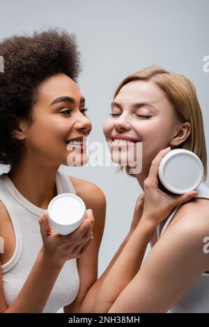 happy blonde model with closed eyes posing with cosmetic cream near african american woman isolated on grey,stock image Stock Photo