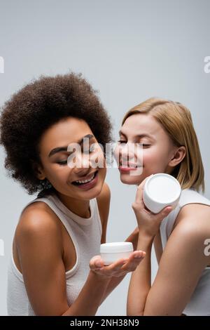 happy african american woman looking at container with body cream near blonde model isolated on grey,stock image Stock Photo