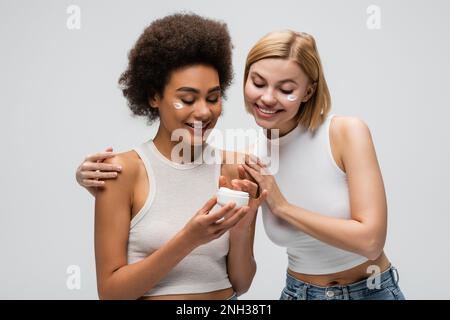 interracial blonde and brunette women smiling near jar of face cream isolated on grey,stock image Stock Photo