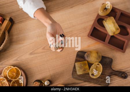 top view of cropped african american woman holding jar with dried herbs near soap bars on chopping board and silicone mold,stock image Stock Photo