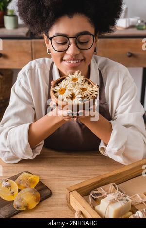 happy african american craftswoman in eyeglasses holding wooden bowl with dried camomiles near bars of handmade soap,stock image Stock Photo