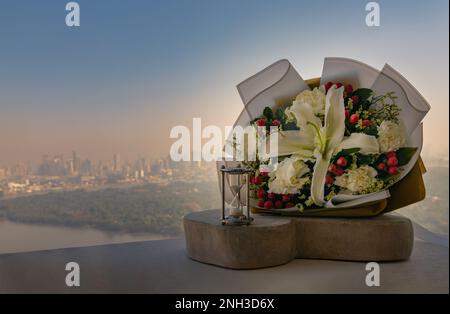 Hourglass or Sandglass is next to Bouquet of flowers placed on the timber outside the balcony with the city view background. Symbol of Romantic foreve Stock Photo