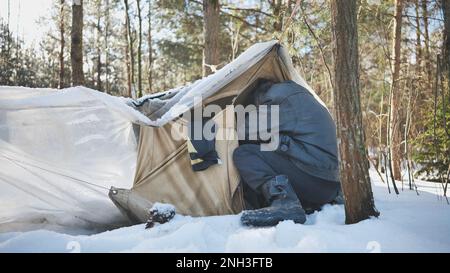 A homeless man climbs in and out of a tent in the woods in winter. Stock Photo