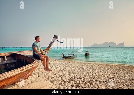 Man sitting on the long tail boat on the sandy tropical beach of Phi Phi island in Southern Thailand. Travel wanderlust magazine concept. Stock Photo