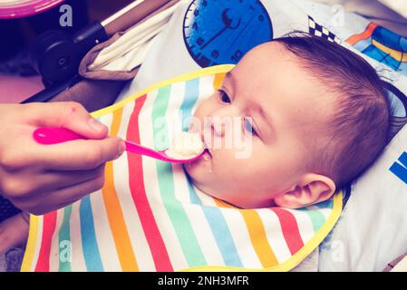 Woman's hand feeding food to little baby boy.New born baby boy wearing napkin while eating cereal. Stock Photo
