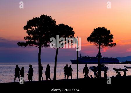 Tourists walking on promenade and cruise ship moored in the Bay of Vlorë, silhouetted against sunset in summer at Vlora in southwestern Albania Stock Photo