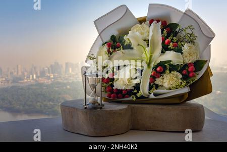 Hourglass or Sandglass is next to Bouquet of flowers placed on the timber outside the balcony with the city view background. Symbol of Romantic foreve Stock Photo