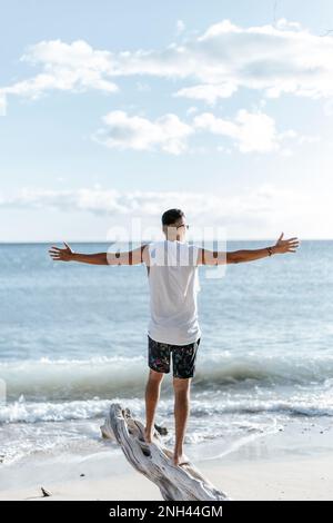 Man on his back with open arms, standing on top of a log, watching the sunset on the beach. Traveling Stock Photo