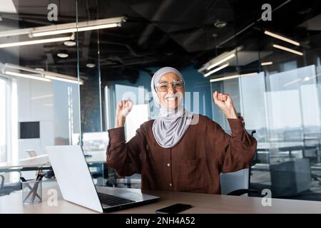 Portrait of happy smiling and successful muslim at work in office, businesswoman with laptop holding hands up gesture of success and triumph, woman in hijab received online victory notification. Stock Photo