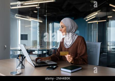 Successful muslim woman in hijab at work inside modern office, business woman with bank credit card and laptop makes online shopping in online store, satisfied and smiling woman at workplace. Stock Photo