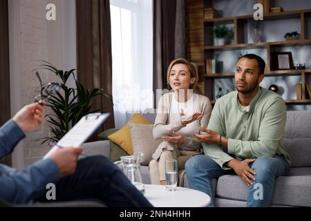 Multiracial quarrel between husband and wife at a session with a psychotherapist. A visit to a family psychologist for a married couple. A doctor Stock Photo
