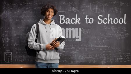 African american male student holding books in front of a blackboard with math formulas and text back to school Stock Photo