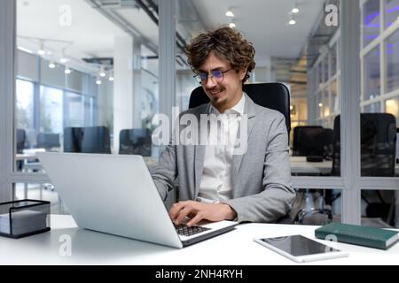 Cheerful and successful businessman inside office working with laptop, bearded man smiling and typing on laptop keyboard, programmer coding software in modern room. Stock Photo