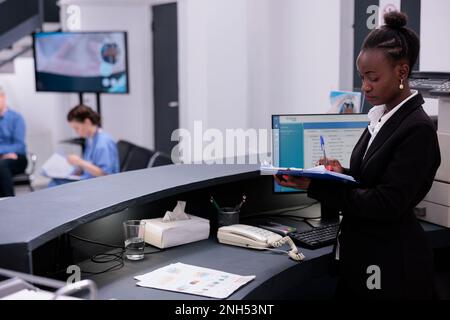 Receptionist in black suit holding clipboard checking medical documents while working at expertise in hospital waiting area. Reception worker is responsible for scheduling appointments Stock Photo