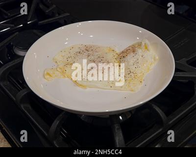 Fried eggs over medium, for breakfast, in a white dinner plate on a stovetop. Stock Photo