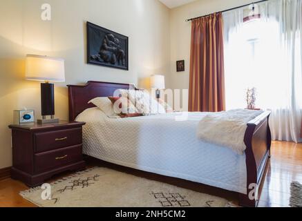 Nightstand and queen size bed with wooden headboard and footboard in guest bedroom on upstairs floor inside luxurious home. Stock Photo