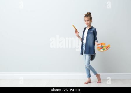Little girl with brush and paint palette near light wall indoors. Space for text Stock Photo