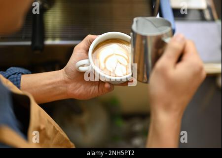 Close-up image, An Asian male barista holding a beautiful latte cup, making beautiful latte art, preparing customer's order in the cafe. Stock Photo