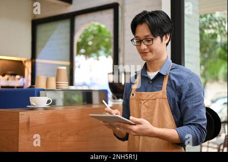 Friendly young Asian male waiter or barista in apron using digital tablet, noting drink orders from a customer in the cafe. small business concept Stock Photo