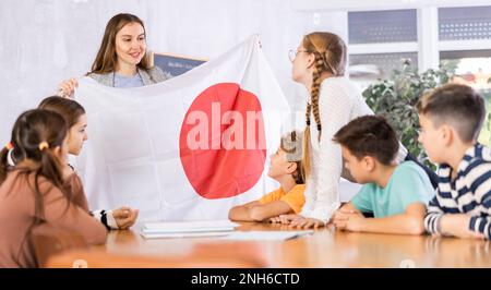 Joyful young woman teacher demonstrating flag for Japan schoolkids preteens during lesson of geography in auditory Stock Photo