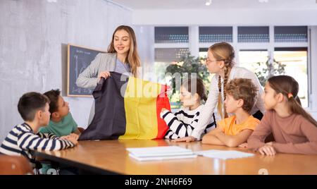 Positive young woman teacher showing flag of Belgium to schoolchildren preteens during history lesson in audience Stock Photo