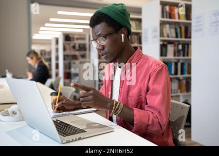 Focused pondered hipster guy in headphones communicates with project team using laptop video call. Stock Photo