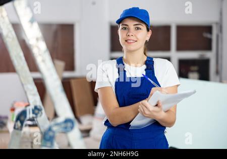 Positive young female builder contractor inspecting room and taking notes Stock Photo
