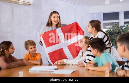 Joyful young woman teacher demonstrating flag of Denmark for schoolkids preteens during lesson of geography in auditory Stock Photo