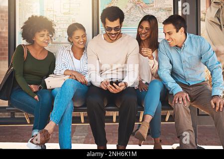 Can everyone see the screen. a cheerful group of young friends seated together while looking on a mobile phones screen outside during the day. Stock Photo