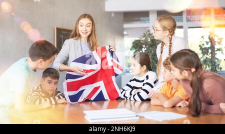 Cheerful young woman teacher demonstrating flag of Great Britain for schoolkids preteens during history lesson in auditory Stock Photo