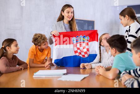 Smiling young woman teacher showing national flag of Croatia and telling preteens schoolchildren history of country during lesson in class Stock Photo