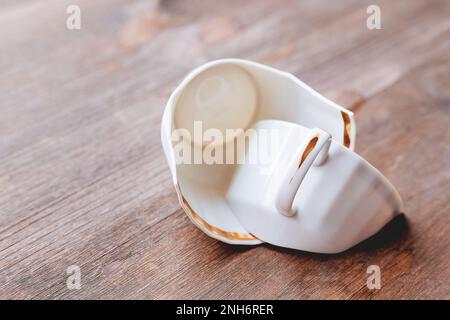 Broken white cup on wooden background. Damaged mug with golden decoration. Stock Photo