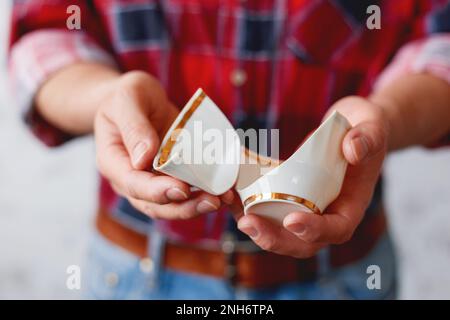 Man in plaid tartan shirt holds a broken white cup. Damaged mug with golden decoration. Stock Photo