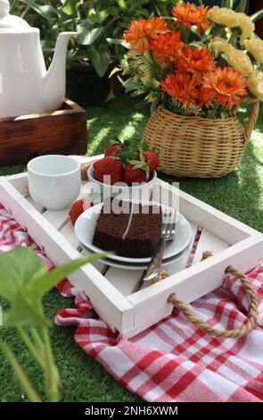 a piece of chocolate cake, a bowl of strawberries and a cup of tea on a wooden tray against a background of flowers in a vase Stock Photo