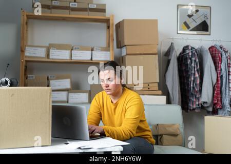 Delivery business. Serious modern female in casual clothes with laptop and parcels using laptop applications in the delivery home office. Copy space Stock Photo