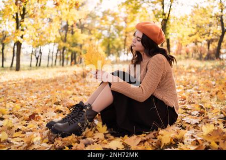 Young brunette Caucasian woman sitting in park in leaves, relaxing on sunny autumn day, holding leaves in her hands. natural lighting, vibrant colors. Stock Photo