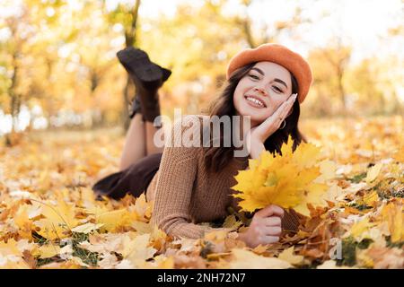Beautiful young woman in park in fall holding leaves. Brunette teenage girl outdoors in autumn lying on the ground. Vibrant colors. Copy space Stock Photo