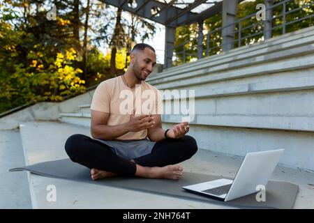 Young African American athlete fitness trainer conducts online training sitting in lotus position, man talking to physical class students using laptop and online video call. Stock Photo