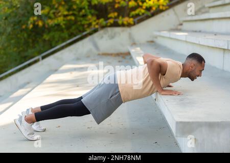 Cheerful and smiling hispanic man doing outdoor exercise in stadium, man stretching and recuperating before jogging in summer park. Stock Photo