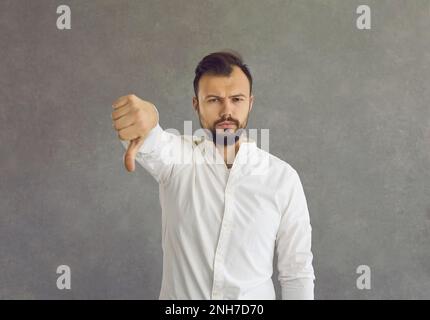Portrait of a young man giving a thumbs down standing isolated on a grey background Stock Photo