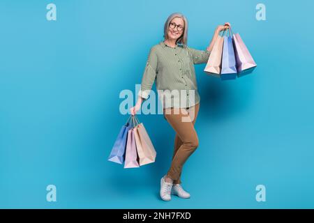 Full length photo of dreamy senior lady dressed striped shirt holding shoppers looking empty space isolated blue color background Stock Photo