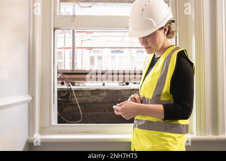 Civil engineer woman using a calculator in front of a window in a construction site, wears yellow high visibility vest, ppe, personal protective equip Stock Photo