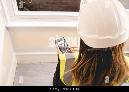 Unrecognizable civil engineer woman with long blond hair calculating with a calculator in a construction site, yellow high visibility vest, personal p Stock Photo